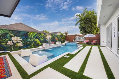 Modern backyard pool with built-in loungers and waterfall, tropical Houston-themed mural on the fence, striped concrete pavers with artificial turf, lounge chairs and umbrella under a bright blue sky