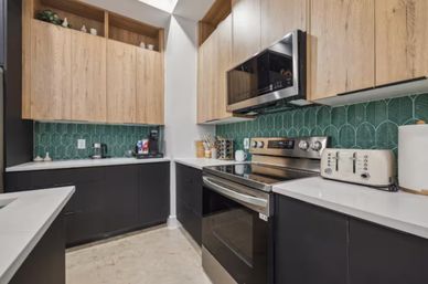 Modern galley kitchen with matte black lower cabinets, light wood upper cabinets, white quartz counters, green scalloped tile backsplash and stainless steel range and microwave with toaster and coffee maker.