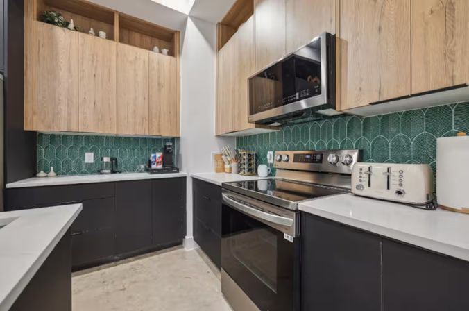 Modern galley kitchen with matte black lower cabinets, light wood upper cabinets, white quartz counters, green scalloped tile backsplash and stainless steel range and microwave with toaster and coffee maker.