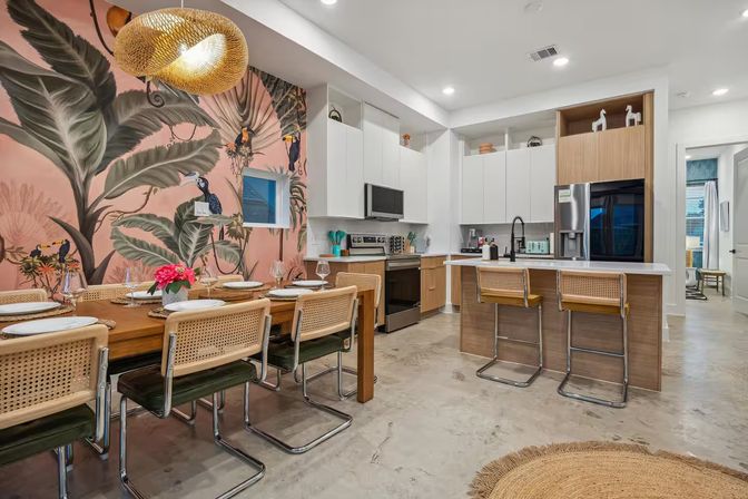 Open-plan modern kitchen and dining area with wooden table, cane-back chairs, breakfast island with barstools, woven pendant light, and a playful pink tropical mural with oversized leaves and toucans.