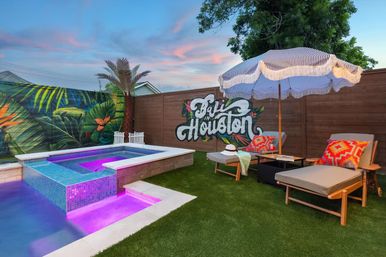 Sunset Houston backyard with purple-lit mosaic spa and pool, tropical mural reading 'Baja in Houston,' lounge chairs with bright pillows, and a striped fringe umbrella on artificial grass.