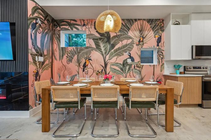 Open-concept dining area with a wooden table and six cane-back chrome chairs, woven pendant light, and a pink tropical mural featuring large palm leaves and toucans, beside a modern kitchen.