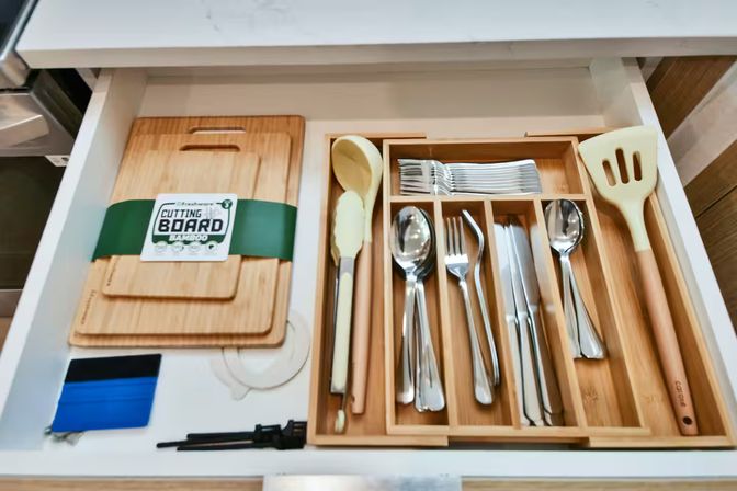 Neatly organized kitchen drawer with stacked bamboo cutting boards and a bamboo cutlery tray holding stainless-steel forks, spoons, knives and wooden and silicone cooking utensils in a modern white drawer.