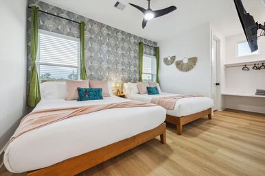 Bright, modern guest bedroom with two queen beds, geometric gray accent wall, green curtains, wood floors, pastel throws and teal tropical pillows, ceiling fan and wall-mounted TV.