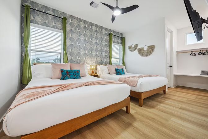 Bright, modern guest bedroom with two queen beds, geometric gray accent wall, green curtains, wood floors, pastel throws and teal tropical pillows, ceiling fan and wall-mounted TV.