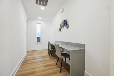 Modern narrow home office nook with built-in gray desk and two gray chairs against white walls, wood-look plank flooring, decorative round wall plates, and a tall slim window letting in natural light.