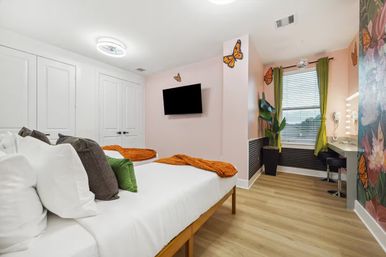 Cheerful butterfly-themed bedroom with two beds, orange throws and green pillows, wall-mounted TV, lighted vanity, potted plant and wood-look floors