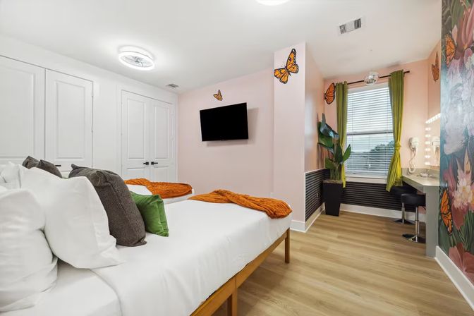Cheerful butterfly-themed bedroom with two beds, orange throws and green pillows, wall-mounted TV, lighted vanity, potted plant and wood-look floors