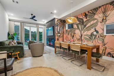 Open-plan modern living and dining room with vibrant tropical mural, long wooden dining table and cane-back chairs, green velvet sofa and round woven pendant light, ceiling fan, concrete floor and sliding glass doors to an outdoor patio.