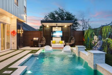 Modern suburban backyard oasis at sunset with illuminated pool, stone water spouts, two in-pool loungers, cabana featuring a wall TV, hanging egg chairs and a sofa with yellow pillows, barbecue grill, tropical mural and seated Buddha statue.