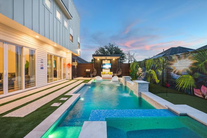 Modern backyard scene at sunset featuring an illuminated rectangular pool with attached spa, two white in-pool loungers, a covered cabana with TV, tropical fence mural, artificial turf walkways and sliding glass doors of a contemporary home