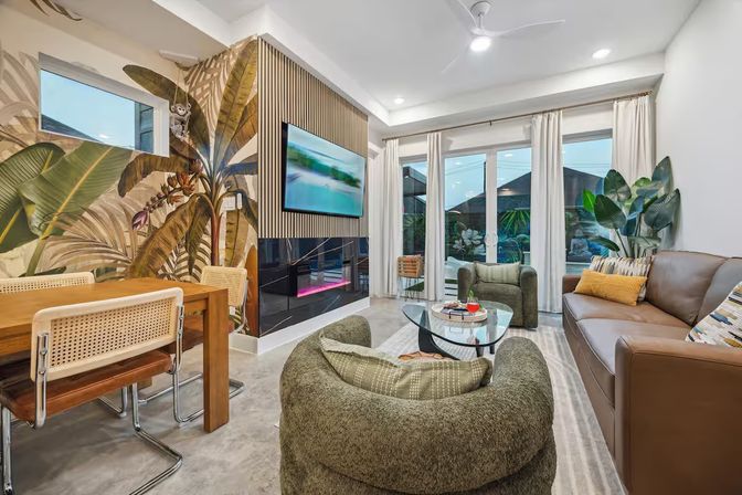 Modern tropical-inspired living room with large banana-leaf mural, wall-mounted TV above a sleek fireplace, plush green swivel chairs, brown leather sofa, wood dining table with cane chairs, glass coffee table, and sliding glass doors to a patio.