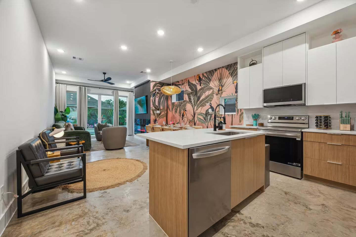 Bright modern open-concept kitchen and living room with wood island, stainless appliances, pink tropical botanical accent wall, pendant light and sliding glass doors to a patio.