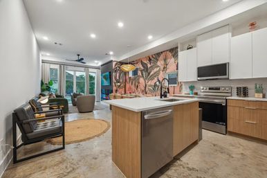 Bright modern open-concept kitchen and living room with wood island, stainless appliances, pink tropical botanical accent wall, pendant light and sliding glass doors to a patio.