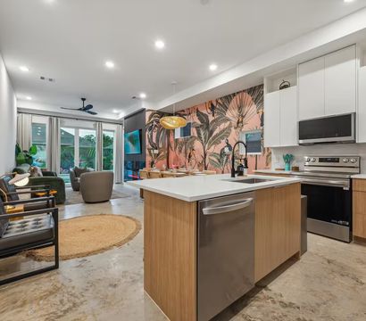 Bright modern open-concept kitchen and living room with wood island, stainless appliances, pink tropical botanical accent wall, pendant light and sliding glass doors to a patio.