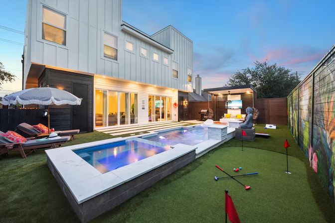 Contemporary two-story home backyard at dusk featuring a lit rectangular pool and spa, artificial-turf putting green with flags, lounge chairs and umbrella, plus a covered outdoor seating area and mural along the fence.
