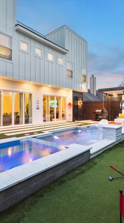 Contemporary two-story home backyard at dusk featuring a lit rectangular pool and spa, artificial-turf putting green with flags, lounge chairs and umbrella, plus a covered outdoor seating area and mural along the fence.