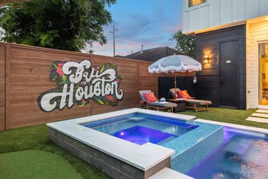 Vibrant Houston backyard at dusk with illuminated blue plunge pool, wooden fence mural reading "Bali in Houston", lounge chairs and a fringed umbrella.
