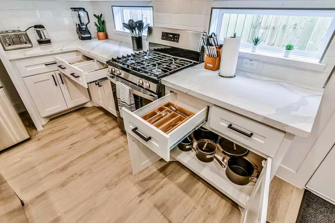 Bright modern white kitchen with stainless steel gas range, marble countertops, open drawers showing a wooden cutlery tray and stacked pots, knife block, small potted succulents on the windowsill, and light wood flooring