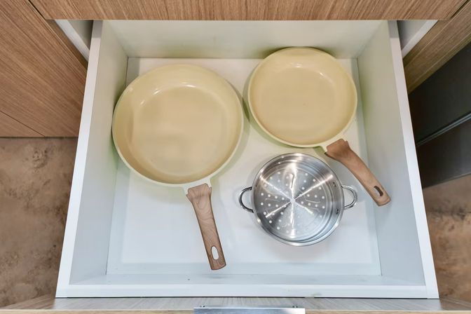 Open kitchen drawer showing two beige frying pans with wood-look handles and a stainless steel steamer insert on a white drawer base.