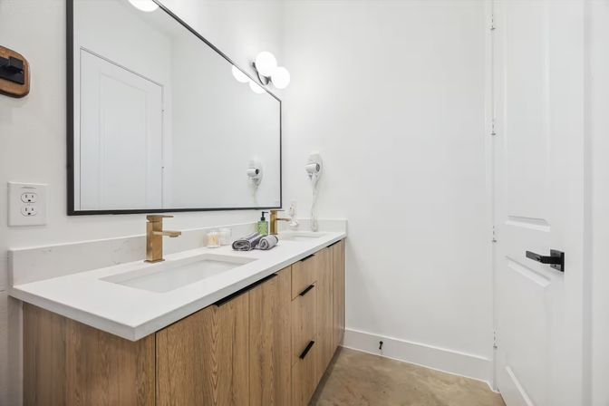 Modern minimalist bathroom with white quartz countertop, oak vanity, gold faucet, large black-framed mirror, wall-mounted hair dryer, folded hand towels and concrete floor