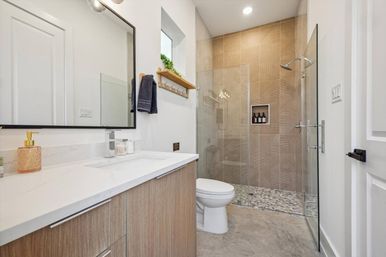 Sleek modern bathroom with wood-grain vanity and white quartz countertop, large mirror, toilet, and frameless glass walk-in shower with textured beige tile, pebble shower floor and recessed niche.