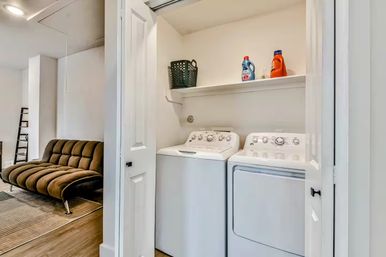 Compact laundry nook in a modern apartment — white top-load washer and matching dryer side-by-side under a shelf with detergent bottles and a basket, open doors revealing a brown tufted futon in the adjacent living area.