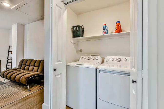 Compact laundry nook in a modern apartment — white top-load washer and matching dryer side-by-side under a shelf with detergent bottles and a basket, open doors revealing a brown tufted futon in the adjacent living area.