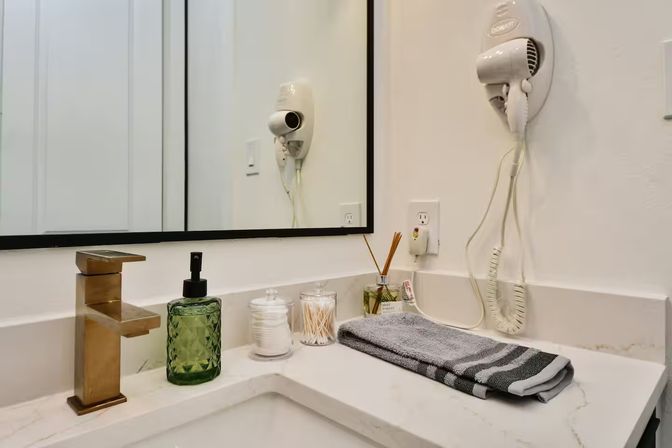Stylish modern bathroom vanity with white marble countertop, gold faucet, green glass soap dispenser, cotton and swab jars, reed diffuser, folded gray towel, wall-mounted hair dryer and mirror.