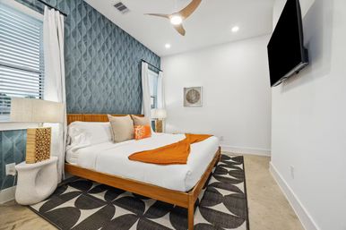 Bright modern coastal bedroom with teal textured accent wall, wooden platform king bed, white linens with orange throw and pillow, patterned black-and-white rug, bedside lamps, ceiling fan and wall-mounted TV.