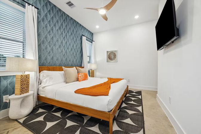 Bright modern coastal bedroom with teal textured accent wall, wooden platform king bed, white linens with orange throw and pillow, patterned black-and-white rug, bedside lamps, ceiling fan and wall-mounted TV.