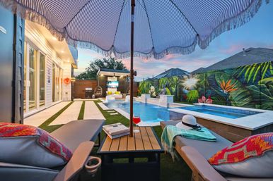 Modern suburban backyard pool and spa at sunset with striped umbrella, lounge chairs, cocktail, towel and hat, cabana with TV and vibrant tropical mural on the fence.