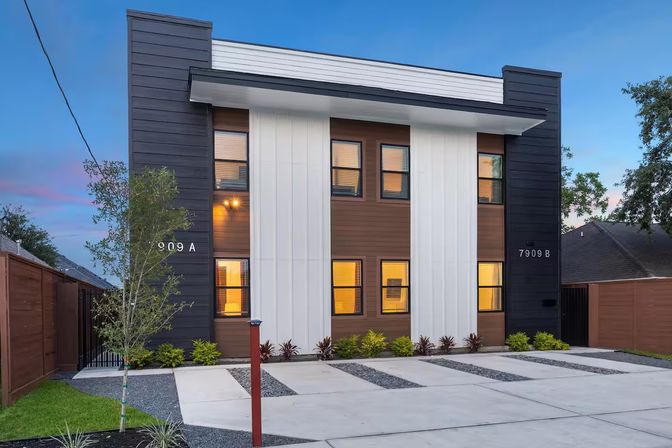 Modern two-story duplex exterior at dusk with dark vertical siding and white central panels, warm-lit windows, concrete driveways with gravel strips, young trees and minimalist landscaping.