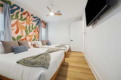 Bright modern guest bedroom with two beds, light wood floors, playful colorful abstract leaf mural on the wall, gray curtains, cozy throw blankets, ceiling fan and wall-mounted TV.