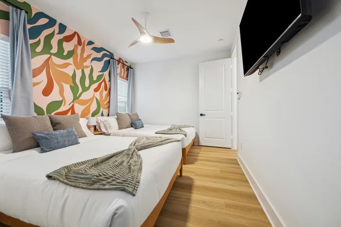Bright modern guest bedroom with two beds, light wood floors, playful colorful abstract leaf mural on the wall, gray curtains, cozy throw blankets, ceiling fan and wall-mounted TV.