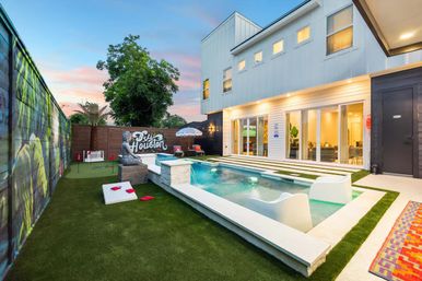 Modern Houston backyard at sunset with a rectangular plunge pool and in-water loungers, artificial turf lawn, cornhole boards, mural-adorned fence, and a two-story white house with large glass doors.
