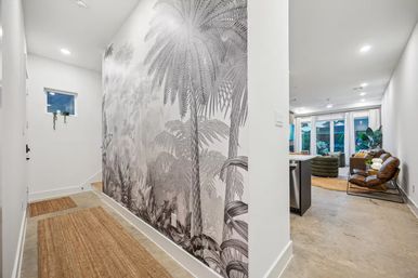Bright modern entry hallway with jute runners and oversized grayscale tropical palm mural opening to an open-concept living room with leather lounge chair, round rug, large windows and indoor plants.