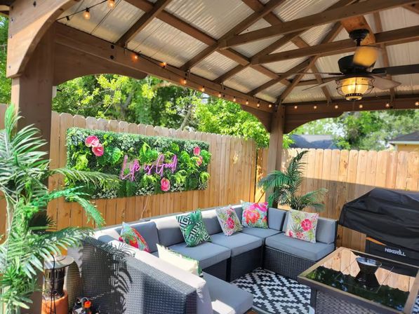 Cozy backyard covered patio with wooden pergola, string lights and ceiling fan, gray wicker sectional with tropical floral pillows, neon “Life is Good” greenery wall and covered grill.