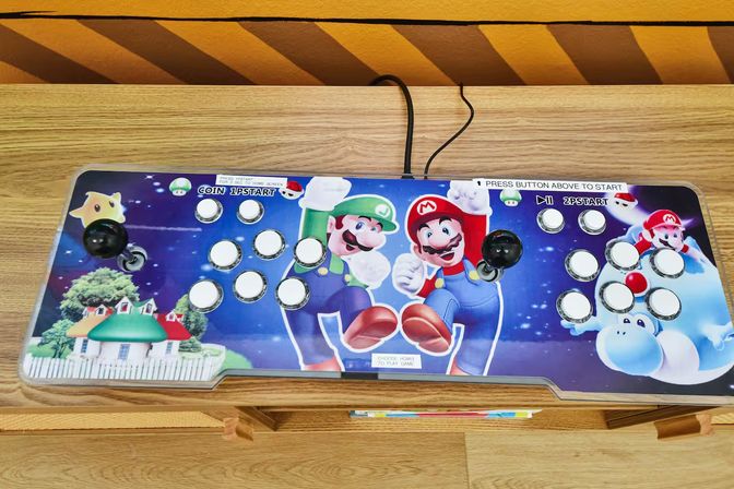 Top-down view of a colorful two-player retro arcade controller with Mario and Luigi artwork, dual black joysticks and multiple glossy white buttons on a wooden table.