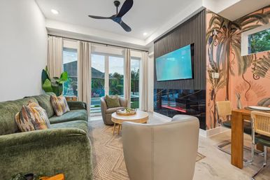 Tropical-inspired modern living room with green velvet sofa and patterned pillows, beige armchairs, round wooden coffee table, wall-mounted TV above a sleek black fireplace, floor-to-ceiling sliding glass doors with pool view, palm-print accent wall, rattan dining chairs and ceiling fan.