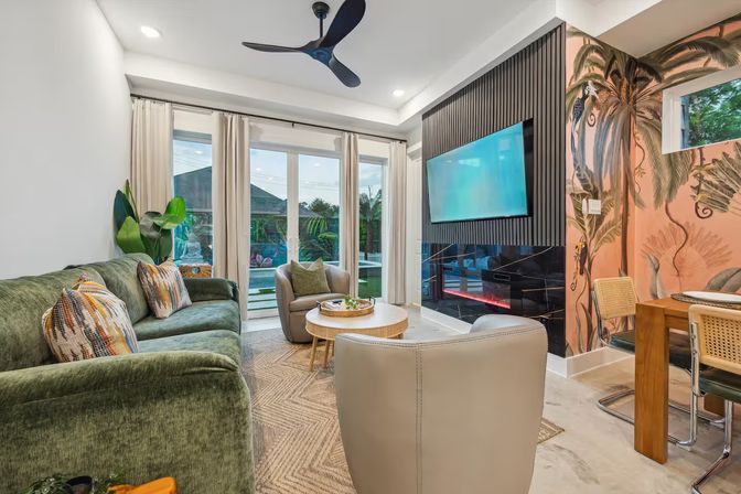 Tropical-inspired modern living room with green velvet sofa and patterned pillows, beige armchairs, round wooden coffee table, wall-mounted TV above a sleek black fireplace, floor-to-ceiling sliding glass doors with pool view, palm-print accent wall, rattan dining chairs and ceiling fan.