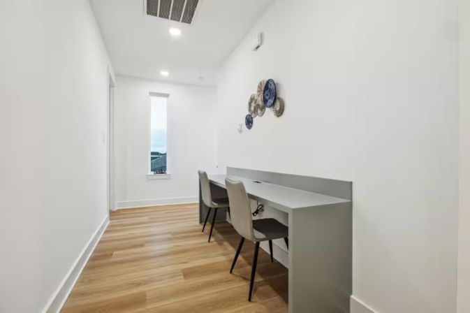 Modern home office nook in a narrow white hallway — built-in gray desk with two chairs, light wood floors, vertical window with natural light, and round decorative wall plates.