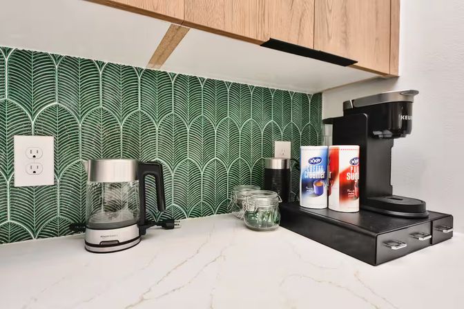 Modern kitchen coffee station on a white marble countertop with a black single‑serve coffee maker, glass electric kettle, small glass jar and creamer and sugar canisters against a green fan‑pattern tile backsplash and light wood cabinets.