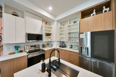 Sleek modern kitchen with white upper cabinets and wood lower cabinets, stainless steel refrigerator and range, open shelving of mugs and dishes, and a white quartz island with a black gooseneck faucet and soap dispensers.