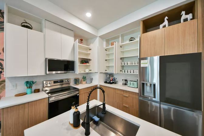 Sleek modern kitchen with white upper cabinets and wood lower cabinets, stainless steel refrigerator and range, open shelving of mugs and dishes, and a white quartz island with a black gooseneck faucet and soap dispensers.