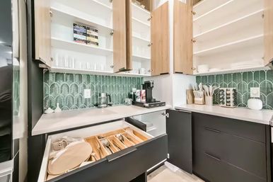 Modern kitchen corner with open light-wood upper cabinets and dark lower drawers, green fan-pattern tile backsplash, white countertop with coffee maker, utensil holders and spice rack, and an open drawer showing a wooden cutlery organizer and plates.