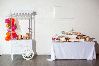 Indoor celebration setup with a white champagne cart decorated with pink and orange balloons and glasses, next to a white-draped dessert buffet of tiered trays, pastries, fresh fruit and floral accents against a white brick wall.