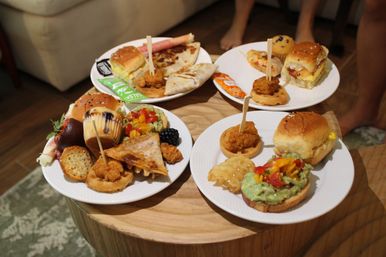 Four plates of assorted brunch finger foods on a round wooden coffee table — mini sliders, chicken-and-waffle bites on skewers, avocado-topped toast, quesadilla wedges, muffins and fruit in a casual indoor setting.