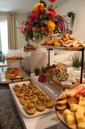 Vibrant home brunch buffet with a tall floral centerpiece and platters of avocado crostini, wraps, pastries, sliders and cheese on a kitchen island.