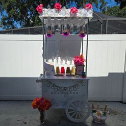 White mobile champagne cart in a sunny backyard, featuring hanging wine glasses and pink disco balls, colorful drink pitchers, ice bucket with bottles, and bright pink floral decorations.
